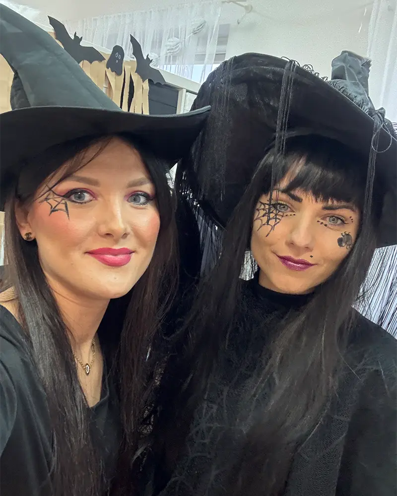 Dwie kobiety w czarnych strojach i kapeluszach czarownic, uśmiechające się do kamery.