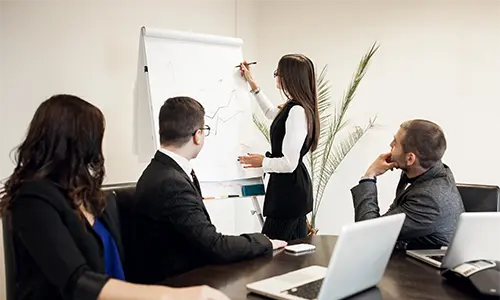 Kobieta prowadzi prezentację dla swoich kolegów w biurze, używając laptopa i gestykulując.