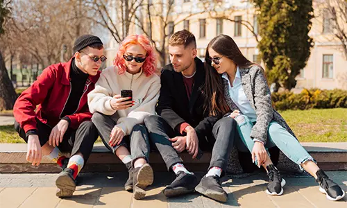 Grupa młodzieży siedząca na murku patrząca się w telefon jednego kolegi.