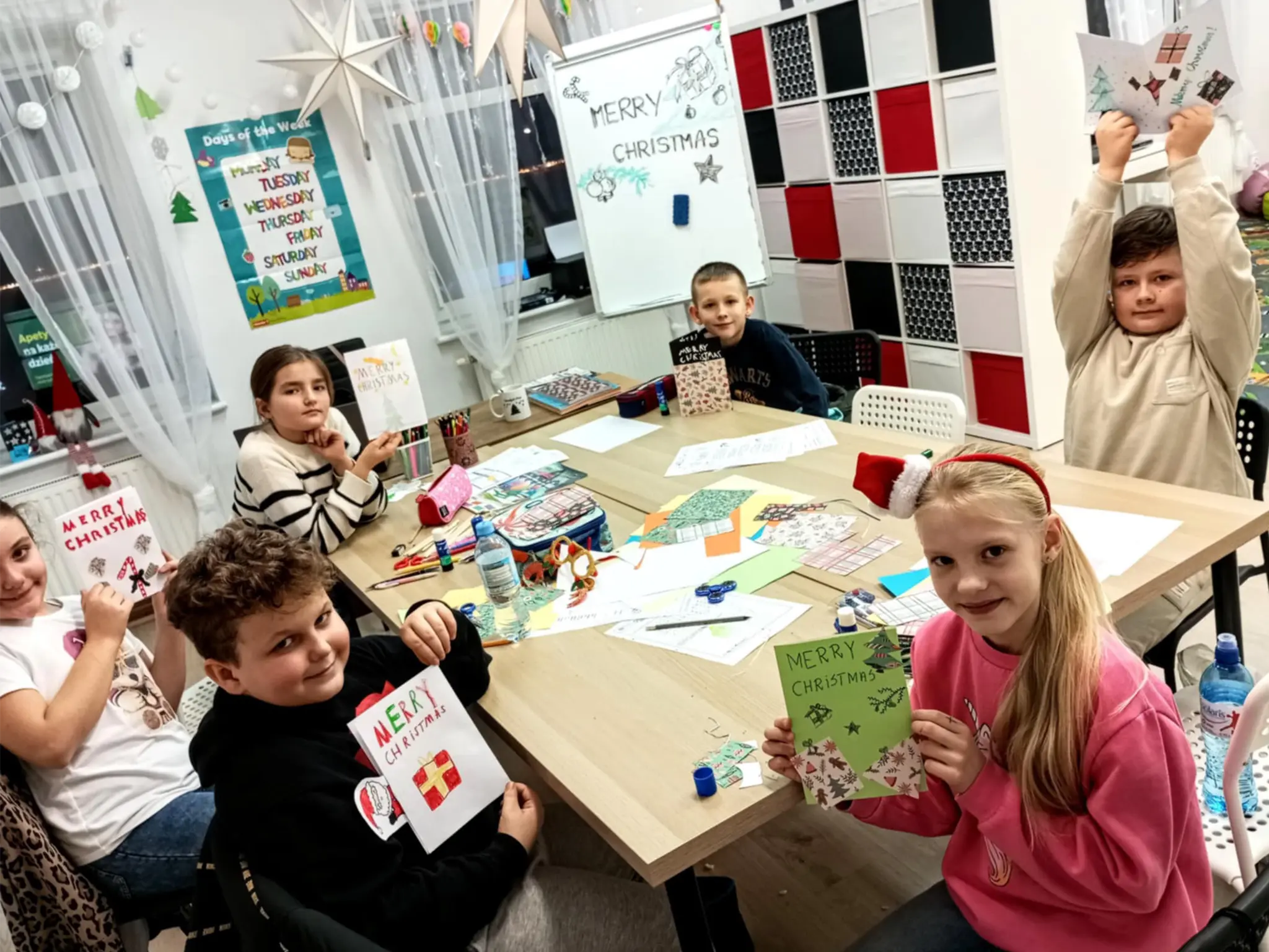 Grupa dzieci przy stole, zajęta grą w karty i tworzeniem rysunków na papierze.