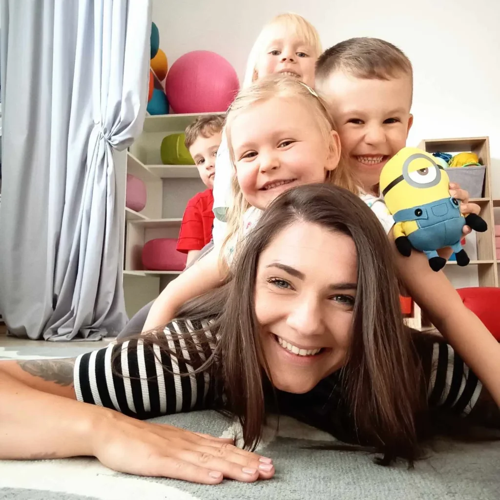 Kobieta leży na podłodze z dziećmi, wszyscy uśmiechnięci i zrelaksowani gotowi na zajęcia językowe.