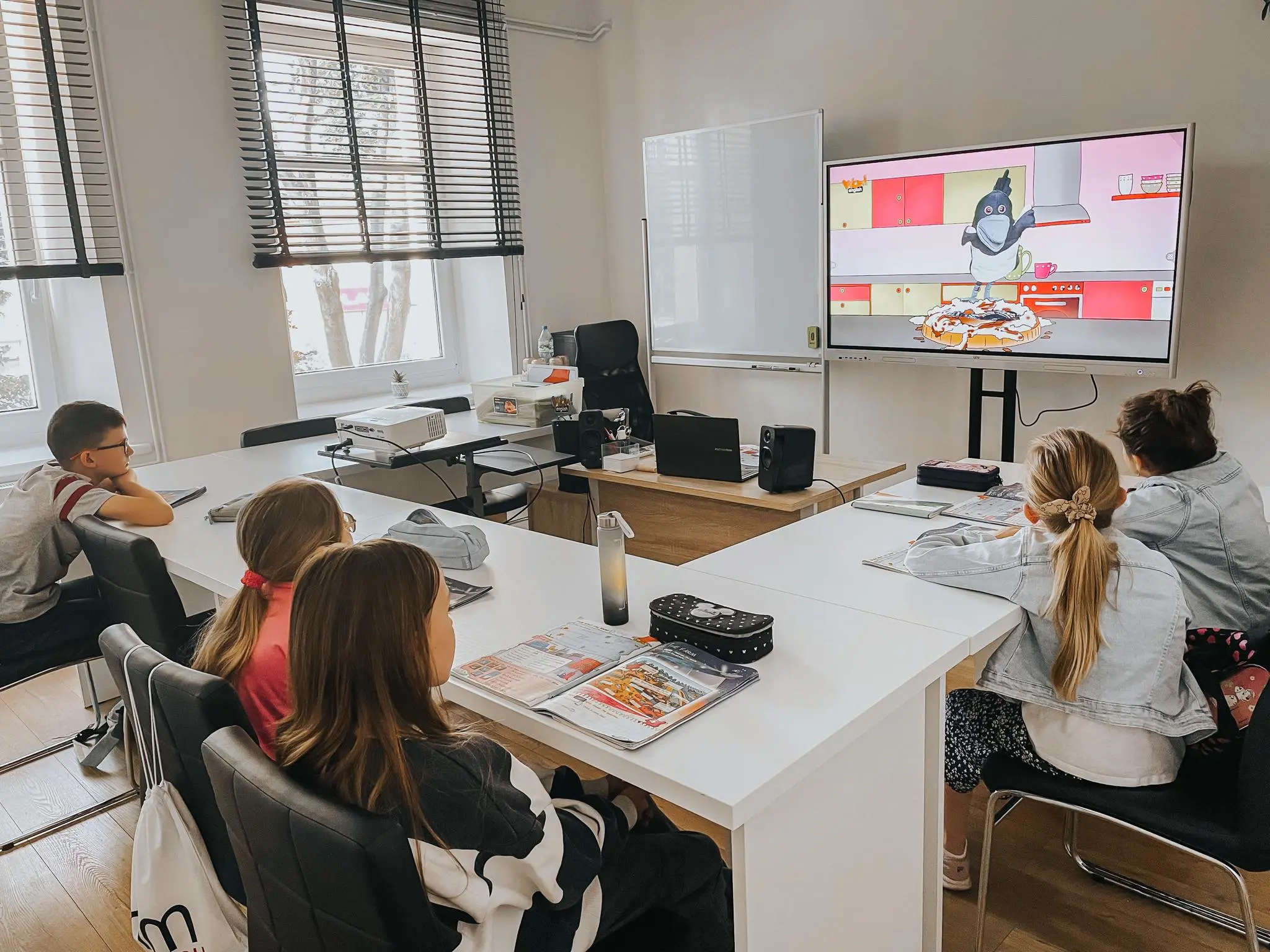 Grupa dzieci siedzi przy biurkach przed telewizorem w sali szkoły językowej.